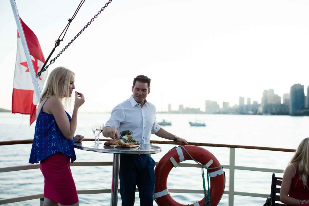 Two people having hor d'oeuvres on boat deck with Toronto in background.
